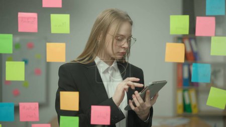 Photo for Brainstorming and ideas in modern business creative concept. Portrait of corporate leader in office planning project on notes. Business woman holding tablet and looking at sticky notes on glass wall. - Royalty Free Image