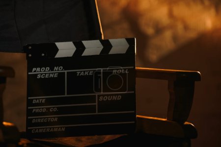 Photo for Movie set and backstage creative advertisement concept. Close up view of black clapperboard standing on directors chair, warm light from projectors on the wall at the background. - Royalty Free Image