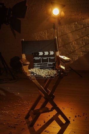 Photo for Movie set and backstage creative advertisement concept. Close up shot cinema clapperboard standing on directors chair popcorn scattered on surface, spotlight with warm light standing at the back. - Royalty Free Image