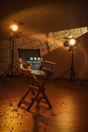 Photo for Movie set and backstage creative advertisement concept. Close up shot of cinema clapperboard standing on directors chair popcorn scattered around, rays of light from the spotlights at the background. - Royalty Free Image