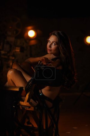Photo for Movie set and backstage creative advertisement concept. Portrait of Beautiful woman actress. Brunette model with makeup in lingerie sitting on directors chair covered by film reels, looking at camera. - Royalty Free Image
