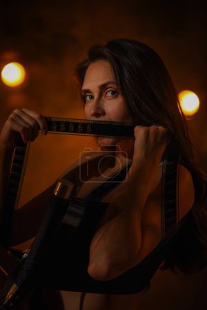 Photo for Movie set and backstage creative advertisement concept. Portrait of Beautiful woman actress. Brunette model with makeup in lingerie sitting on directors chair covering mouth with film reels, close up. - Royalty Free Image
