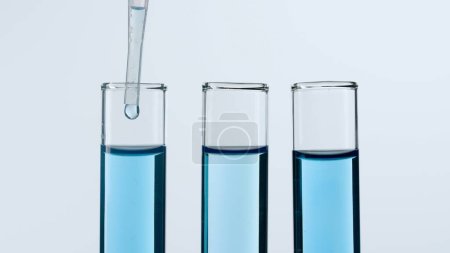 Foto de Tres tubos de ensayo de vidrio sobre fondo blanco. Dos tubos de ensayo están llenos de líquido azul, una sustancia azul gotea en uno de ellos desde una pipeta. Concepto de medicina, investigación bioquímica. Cierre - Imagen libre de derechos