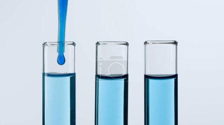 Foto de Tres tubos de ensayo de vidrio sobre fondo blanco. Los tubos de ensayo están llenos de líquido azul, una sustancia azul gotea en uno de ellos desde una pipeta. Concepto de medicina, investigación bioquímica. Primer plano - Imagen libre de derechos