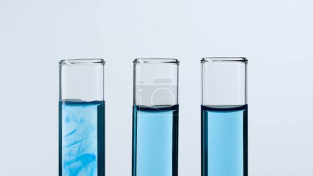 Foto de Tres tubos de ensayo de vidrio sobre fondo blanco. Los tubos de ensayo están llenos de líquido azul, uno muestra una sustancia disolvente azul. Concepto de medicina, investigación bioquímica. Primer plano - Imagen libre de derechos