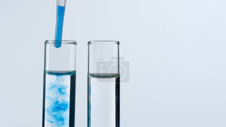 Foto de Dos tubos de ensayo de vidrio sobre fondo blanco. Los tubos de ensayo están llenos de líquido transparente, una sustancia azul gotea en uno de ellos desde una pipeta. Concepto de medicina, investigación bioquímica - Imagen libre de derechos
