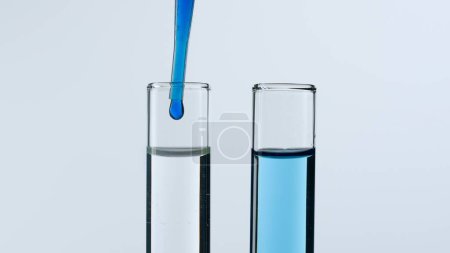 Foto de Dos tubos de ensayo de vidrio sobre fondo blanco. Los tubos de ensayo están llenos de líquido, una sustancia azul gotea en uno de ellos desde una pipeta. Concepto de medicina, investigación bioquímica. Primer plano - Imagen libre de derechos