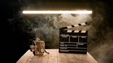 Photo for A clapper board and vintage video camera on a wooden table enveloped in smoke. Cinematography equipment in a studio on a black background illuminated by a neon lamp - Royalty Free Image