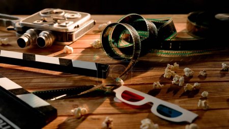 Téléchargez les photos : Clapper board, caméra vidéo vintage, film et verres 3D avec grains de maïs soufflé sur table en bois dans des nuages de fumée. Concept de cinématographie - en image libre de droit
