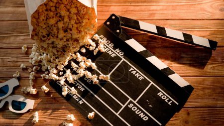 Photo for Fresh salty popcorn from a paper bag is scattered on a movie clapper board. Popcorn, movie clapper board and 3d glasses on wooden table. Cinema for two concept - Royalty Free Image