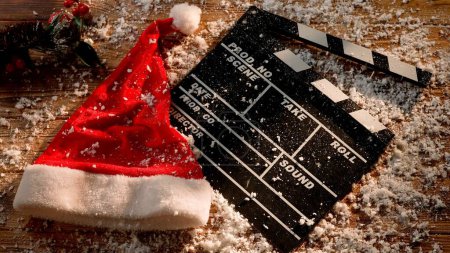 Téléchargez les photos : La neige recouvre une planche de film, un chapeau de Noël et une branche d'épinette sur une table en bois. Un concept basé sur la sortie de films de vacances de Noël et de productions cinématographiques - en image libre de droit