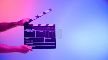 Foto de La mano sostiene una pizarra negra sobre un fondo azul con una luz circular, iluminada con luz de neón rosa. Industria cinematográfica, entretenimiento - Imagen libre de derechos