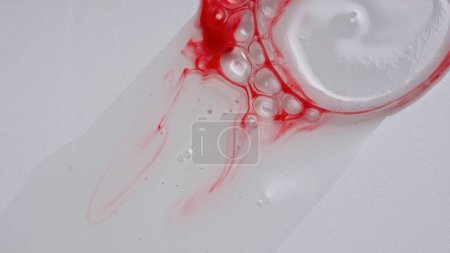 Foto de Concepto de publicidad creativa en gel y líquido. Primer plano de sustancia transparente sobre el fondo blanco. Burbuja grande con flujos de tinta roja sobre el producto cosmético gel fluido o suero en la superficie. - Imagen libre de derechos