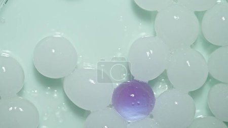 Téléchargez les photos : Beaucoup de sphères d'hydrogel blanc avec une violette en lumière verte. Les sphères rondes brillantes de gel scintillent sur le fond blanc du studio - en image libre de droit