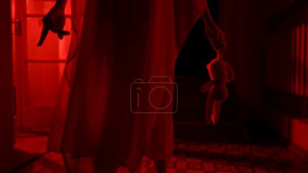 Photo for Horror movie and poltergeist creative advertising concept. Female ghost in a house. A woman in a white dress is walking down a hallway holding a toy. Red horror light. Close up - Royalty Free Image