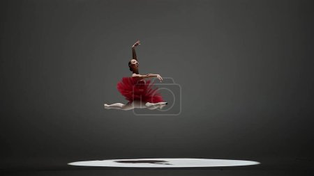 Photo for A ballerina in a red tutu and pointe shoes during a twine jump. A young woman dancing elements of classical ballet in a dark studio with overhead light - Royalty Free Image