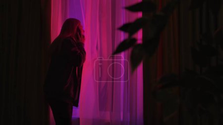Photo for Woman talking on smartphone and looking out window at red and blue police lights. Curious woman in a dark apartment looking out the window - Royalty Free Image