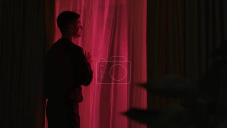 Photo for A curious, concerned man looks out the window at the red lights of an emergency service, police, ambulance or fire department. The man is curious about what has happened - Royalty Free Image