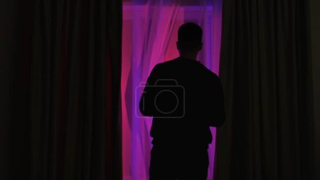 Photo for Back view of a man standing near a window in a dark room looking out into the street. Man looking at lights of police or other emergency service - Royalty Free Image