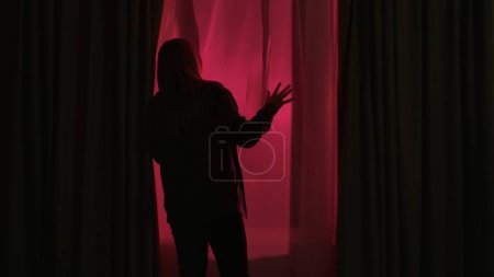 Photo for Back view of a curious woman standing near a window in a dark room looking out into the street. Woman looking at lights of police or other emergency service - Royalty Free Image