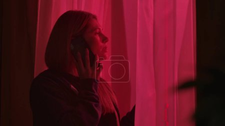Foto de Una mujer de pie cerca de una ventana en un cuarto oscuro iluminado por luces de la policía, de cerca. Mujer preocupada hablando en un smartphone - Imagen libre de derechos