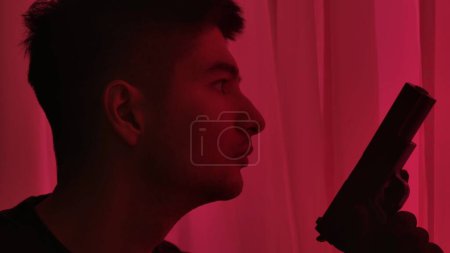 Téléchargez les photos : Portrait d'un criminel près d'une fenêtre éclairée par la lumière rouge provenant des sirènes de la police. Homme criminel tenant une arme et regardant par la fenêtre - en image libre de droit