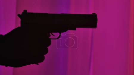 Foto de Un arma en la mano de un criminal, contra el fondo de una ventana iluminada por luces de la policía, de cerca - Imagen libre de derechos