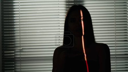 Photo for Beauty of body creative advertisement concept. Portrait of female model in studio. Appealing woman silhouette with light pointing on part of the body and face standing against window with jalousie. - Royalty Free Image