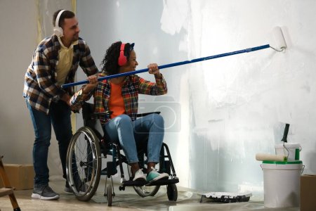 Photo for Depicted is a room in which a renovation is taking place. A couple is making repairs, painting the walls. The woman is in a wheelchair, the man is helping her. They are wearing headphones and smiling. - Royalty Free Image