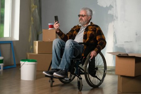 Photo for The shot shows a room in which a renovation is taking place, it is unfinished. In the center of the room sits a man in a wheelchair, he stares intently into his phone. General plan. - Royalty Free Image