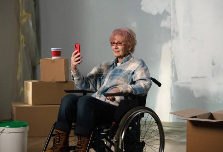 Photo for The shot shows a room in which a renovation is taking place, it is unfinished. In the center of the room sits a woman in a wheelchair, she is stares intently into her phone. General plan - Royalty Free Image