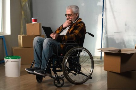 Photo for The shot shows a room in which a renovation is taking place, it is unfinished. In the center of the room sits a man in a wheelchair, he is stares intently into his tablet. General plan. - Royalty Free Image