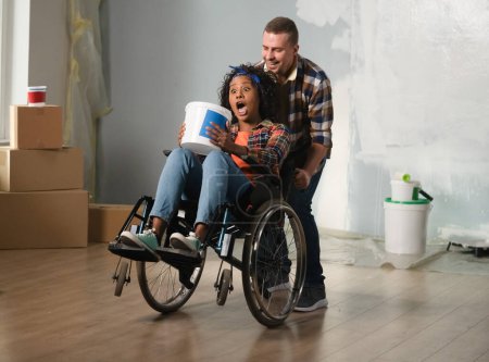 Photo for The shot shows a room being renovated. In the center stands a man and a woman in a wheelchair. She is holding a bucket of paint and he has lifted her up, they are happy, laughing. General outline. - Royalty Free Image