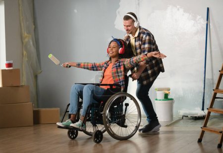 Photo for The shot shows a room being renovated. In the center stands a man and a woman in a wheelchair. They are wearing headphones having fun, enjoying themselves, happy and laughing. General outline. - Royalty Free Image