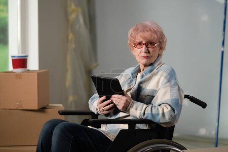 Photo for The shot shows a room that is being renovated, it is not yet finished. In the center of the room sits a woman in a wheelchair. She is holding a tablet. General plan. - Royalty Free Image
