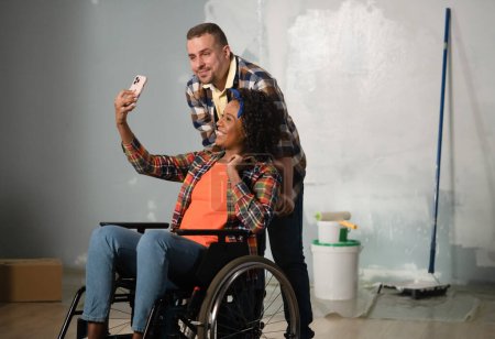 Photo for The shot shows a room that is being renovated, it is not yet finished. In the center of the room is a man and a woman in a wheelchair. They are taking selfies, posing for the camera. - Royalty Free Image