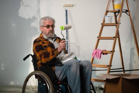 Photo for The image shows a room in which renovations are underway, they are not yet finished. In the center of the room sits a man in a wheelchair. He is holding a paint roller and scrutinizing. - Royalty Free Image