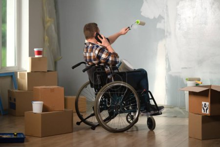 Photo for The image shows a room in which renovations are underway, they are not yet finished. In the center of the room sits a man in a wheelchair. He is talking on the phone and painting the walls. - Royalty Free Image