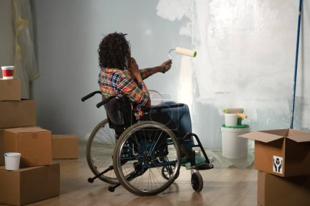 Photo for The image shows a room in which renovations are underway, they are not yet finished. In the center of the room sits a woman in a wheelchair. She is talking on the phone and painting the walls - Royalty Free Image