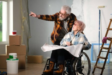 Photo for A room being renovated is shown. A woman in a wheelchair, next to her stands a man, they are elderly and shows somewhere, she is holding a drawing of a house. They are happy, smiling, dreaming. - Royalty Free Image