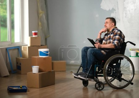 Photo for The shot shows a room in which renovation is in progress, it is not yet finished. In the center of the room sits an adult man in a wheelchair. He has a tablet in his hands, pensive. - Royalty Free Image