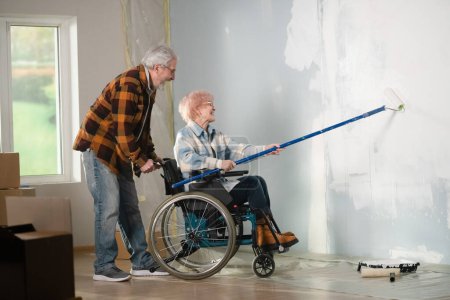 Photo for The shot shows a room in which a renovation is taking place. A couple is making repairs, painting the walls with white paint. The woman is in a wheelchair, the man is helping her. They are attentive. - Royalty Free Image