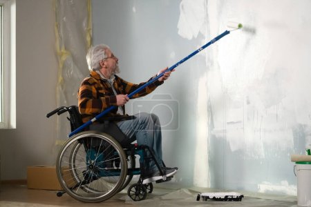Photo for The picture shows the room in which the repair is carried out. An elderly man in a wheelchair. He is a repairman who paints a roller on a long stick of paint on the wall. - Royalty Free Image