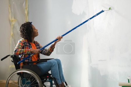 Photo for The picture shows the room in which the repair is done. An adult woman in a wheelchair. She is engaged in repair painting roller on a long stick wall white paint. - Royalty Free Image