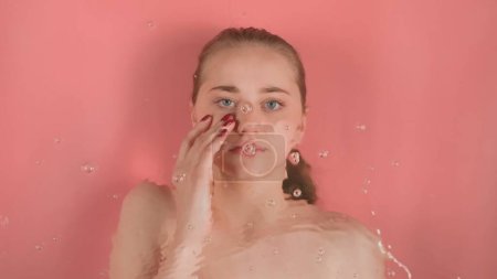 Photo for Young girl with bare shoulders without makeup on pink background. She is lying underwater, looking at the camera and touching her face with her red manicured fingers. Waves form on the water - Royalty Free Image