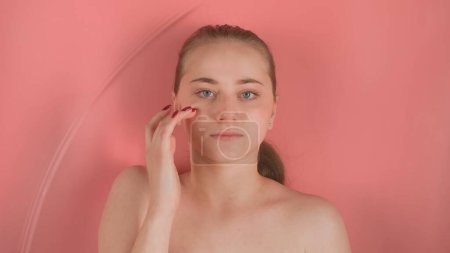 Téléchargez les photos : Jeune fille aux épaules nues sans maquillage sur fond rose. Elle est allongée sous l'eau, regarde la caméra et touche son visage avec ses doigts rouges manucurés. Des vagues se forment sur l'eau - en image libre de droit