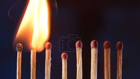 Foto de Una fila de fósforos donde dos fósforos en llamas pasan su fuego al siguiente. Cascada de llama, transferencia de energía. Macro disparo de la transición de la llama de un partido a otro - Imagen libre de derechos