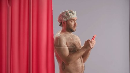 Photo for Man in waterproof shower cap in studio on blue background using smartphone. Man standing near pink shower curtain with a dreamy look typing a message, browsing social media, smiling sending an air - Royalty Free Image
