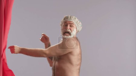 Foto de Un hombre con espuma de afeitar en la barba se asoma por detrás de una cortina de ducha rosa y grita asustado. Varón con torso desnudo y gorra de ducha. Concepto de belleza humorístico - Imagen libre de derechos