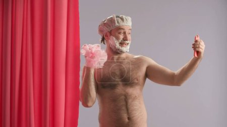 Téléchargez les photos : Homme avec mousse à raser sur sa barbe prendre des selfies avec loofah à l'aide d'un smartphone. Un homme au torse nu et coiffé d'un bonnet de douche. Concept de beauté humoristique - en image libre de droit